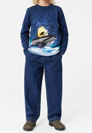 Lapsi, joka pitää yllä tummansinistä pitkähihaista paitaa, jossa on avaruusteleskoopin grafiikka, löysi sinisiä farkkuja ja ruskeita kenkiä, seisoo kädet taskuissa.