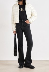 Puffig creamfärgad jacka med hög krage, svart T-shirt med guldtryck, svarta utsvängda jeans och svarta skor, håller en svart väska.