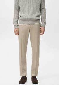 Pantalon beige à coupe ajustée, avec des plis frontaux nets et des poches latérales, associé à un pull gris clair côtelé.