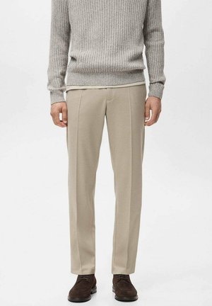 Pantalones de vestir beige con un corte entallado, con pliegues delanteros marcados y bolsillos laterales, combinados con un suéter gris claro de tejido acanalado.