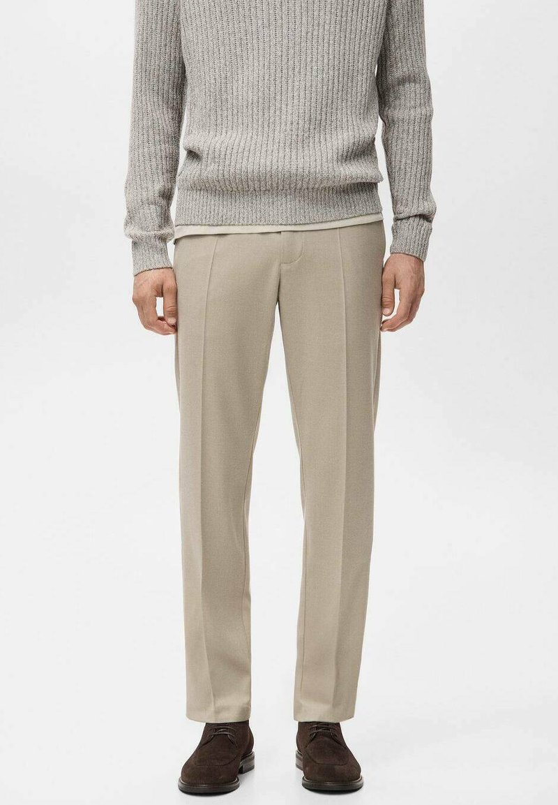 Pantalon beige à coupe ajustée, avec des plis frontaux nets et des poches latérales, associé à un pull gris clair côtelé.