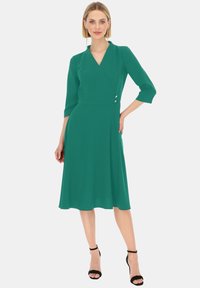 Turkusowa sukienka zabudowana z trzema ćwierć rękawami, dekoltem w kształcie litery V, dopasowaną talią, rozkloszowanym dołem oraz dwiema srebrnymi guzikami na boku.