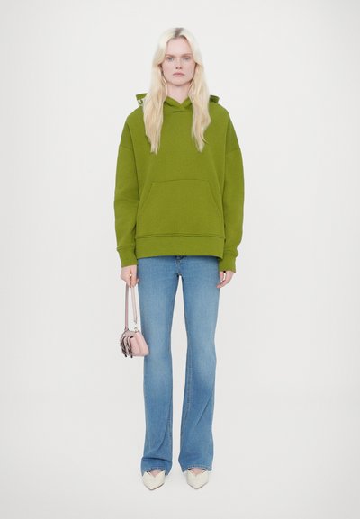 Grön huvtröja med ficka fram, kombinerad med blå utsvängda jeans. Modellen håller en liten rosa handväska och bär spetsiga vita skor.