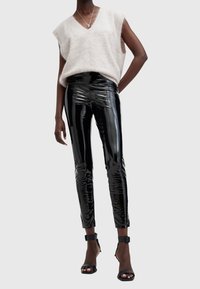 Mannequin portant un haut beige sans manches duveteux, un pantalon en vinyle noir brillant taille haute, et des sandales noires à talons hauts avec bride à la cheville.