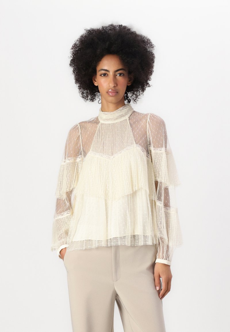 Light cream blouse with sheer fabric, textured ruffles, and polka dots. High collar and long sleeves. Paired with light beige pants.