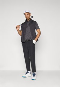 Man in black golf vest and pants, white cap and shoes, holding golf club over shoulder, standing against plain gray background.