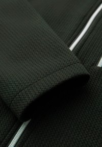 Mörkgrön texturerad tyg med ett bikakesmönster, med vita dragkedjor längs sömmarna. Synlig ärmdetalj.