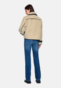 Beige faux fur jacket with black accents, cropped design, and a pointed collar, paired with blue denim flared jeans and black boots.