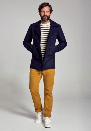 Homme barbu portant un caban bleu marine, une chemise rayée, un pantalon moutarde et des baskets blanches, debout les mains dans les poches devant un fond uni.
