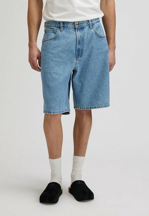 Uomo che indossa pantaloncini di denim azzurro chiaro con una t-shirt bianca, calzini bianchi e scarpe slip-on nere, in piedi davanti a uno sfondo neutro.