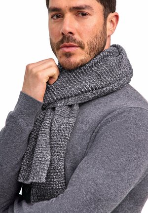 Mężczyzna z krótkimi ciemnymi włosami i brodą, poprawiający szary, masywny dziergany szalik, ubrany w pasujący szary sweter, na białym tle.