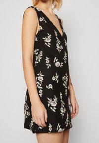 Vestido negro sin mangas con escote en V profundo, que presenta bordados florales blancos. La tela tiene un acabado texturizado y una silueta ajustada.