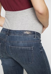 Top ajustado gris y marrón combinado con jeans de mezclilla azul oscuro que cuentan con un bolsillo trasero y una etiqueta de marca en la cinturilla.