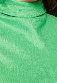 Zielony golf wykonany z teksturowanej tkaniny, cechujący się gładkim wykończeniem i wysokim kołnierzem. Kolor jest jasny i żywy.