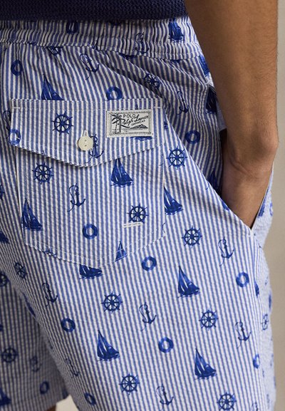 Μαγιό βερμούδα με μπλε και λευκές ρίγες, ναυτικά εικονίδια σε σκούρο μπλε και πίσω τσέπη με κουμπί, φορεμένο από άτομο με το χέρι στην πλαϊνή τσέπη.