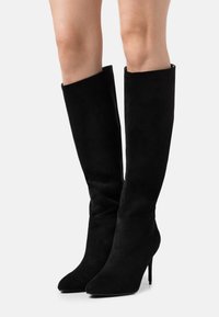 Bottes noires montant jusqu'aux genoux en suède doux. Dotées d'un bout pointu et d'un talon stiletto. Texture lisse sans motifs ni accents visibles.