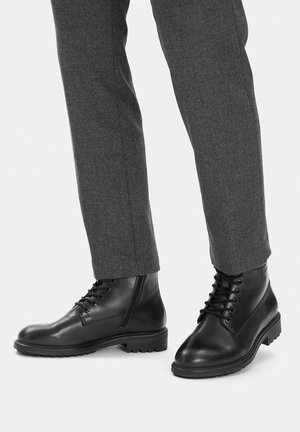 Schwarze Leder-Schnürstiefel, getragen zu grauen, gerade geschnittenen Hosen an einer stehenden Person vor hellem Hintergrund.