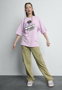 T-shirt oversized lilas avec un graphique de palmier, associé à un pantalon cargo olive ample et des baskets grises avec des accents blancs.