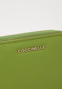Zielona skórzana portmonetka o teksturowanej powierzchni, ze złotymi elementami i zamkiem błyskawicznym. Posiada wytłoczone logo "Coccinelle" z przodu.