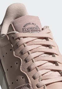 Zapatilla de cuero rosa claro con lengua texturizada, cordones de tela, lados perforados y un parche circular de logo en la lengua.