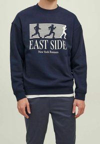 Uomo che indossa una felpa blu navy con figure bianche di corridori e la scritta "EAST SIDE New York Runners" sulla parte anteriore.