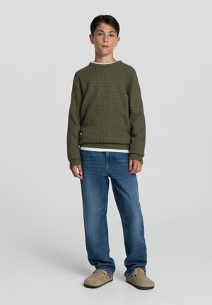 Maglione a trama verde oliva sopra uno strato bianco, abbinato a jeans in denim blu e scarpe sportive beige. Design semplice e vestibilità regolare.