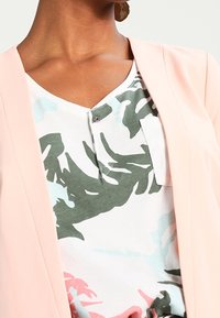 Blush pink blazer layered over a white shirt with tropical green and light blue leaf patterns; features a buttoned v-neck.