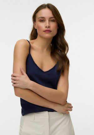 Nő hosszú barna hajjal, sötétkék ujjatlan felsőt és világos bézs nadrágot viselve, karját összefonva áll, egyszerű háttér előtt.