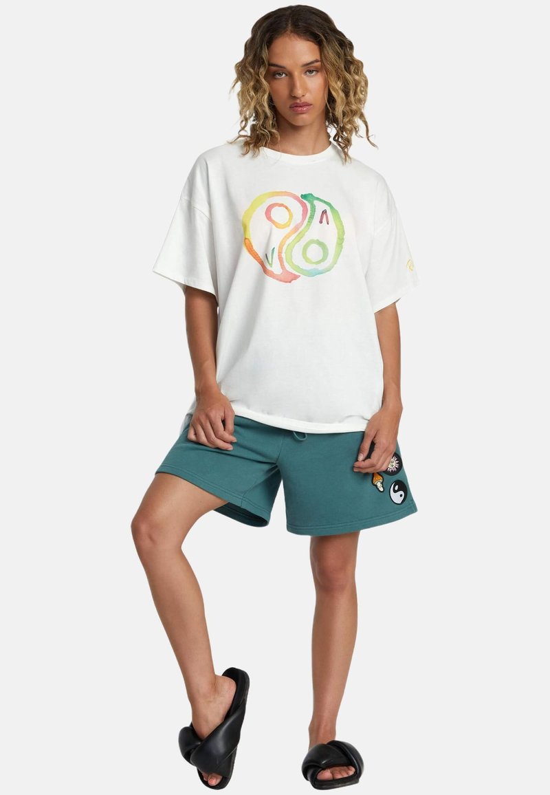 Camiseta blanca de gran tamaño con un gráfico colorido de yin-yang, combinada con pantalones cortos en color teal y sandalias negras de tipo deslizante. Los pantalones cortos cuentan con parches decorativos.