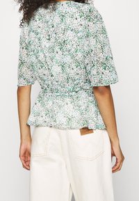 Blusa peplum floreale in tessuto leggero e trasparente con motivi verdi, bianchi e viola; presenta maniche corte e vita arricciata.