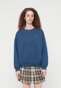 Μπλε φούτερ με στρογγυλή λαιμόκοψη, μακριά μανίκια και διακριτικό γραφικό στο στήθος. Συνδυασμένο με καρό, πλισέ φούστα.