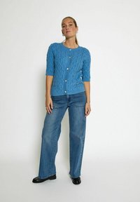 Blå kabelstickad cardigan med korta ärmar, framknäppning och ett strukturerat mönster, tillsammans med ljusblå vida jeans och svarta skor.