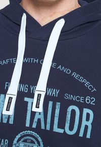 Felpa blu navy con cappuccio e lacci bianchi, caratterizzata da testo blu stampato e accenti di logo. Texture morbida, design casual e stile con cappuccio.