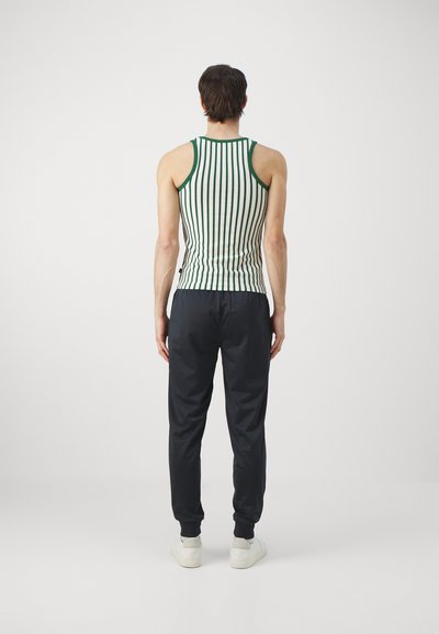 Stripet tanktop med grønne og hvite vertikale striper; kombinert med marineblå joggebukser; modellen har på seg hvite sneakers. Avslappet passform, minimal design.