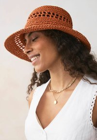Sombrero de paja trenzada marrón con ala ancha y detalles recortados, complementado con collares de oro en capas y una camiseta sin mangas blanca.
