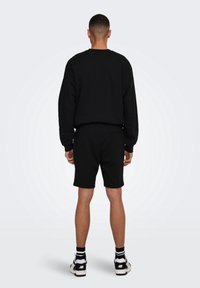 Uomo di spalle che indossa una maglietta nera a maniche lunghe, pantaloni corti neri, calze a righe e scarpe da ginnastica bianche e nere su uno sfondo bianco uniforme.