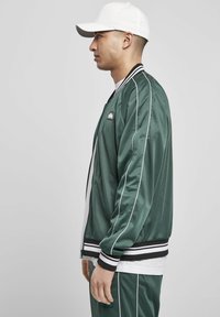Giacca bomber in satin verde con strisce bianche, polsini neri a coste e una toppa con logo. Indossata sopra una t-shirt bianca, abbinata a pantaloni coordinati.