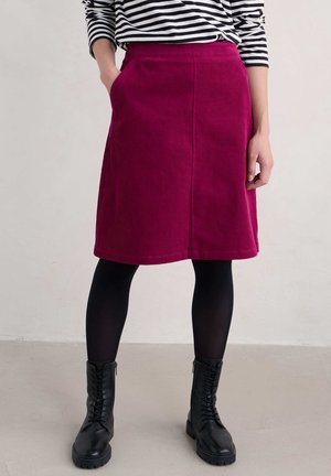 Frau trägt einen fuchsienfarbenen Cord-Knielangrock, schwarze Strumpfhosen, schwarze Schnürstiefel und ein schwarz-weiß gestreiftes Langarmshirt.