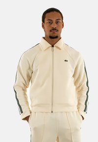 Chaqueta de color beige claro con cremallera, hecha de un material suave, que presenta franjas laterales verdes y un pequeño logo bordado en el lado izquierdo del pecho.
