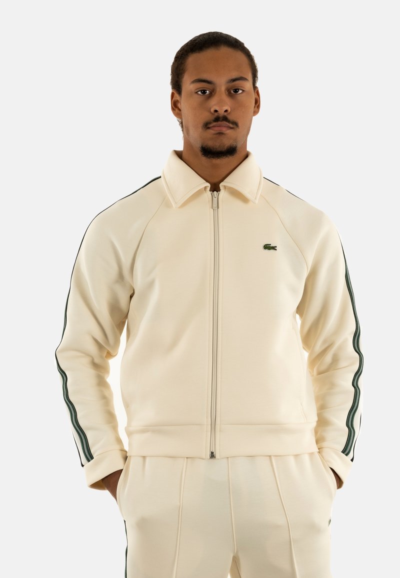 Chaqueta de color beige claro con cremallera, hecha de un material suave, que presenta franjas laterales verdes y un pequeño logo bordado en el lado izquierdo del pecho.