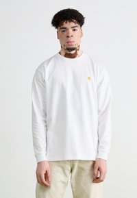 Chemise blanche à manches longues en coton avec un col rond. Présente un petit logo jaune sur le côté gauche de la poitrine. Texture lisse.