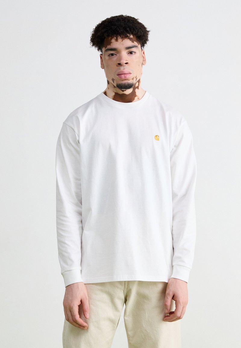 Chemise blanche à manches longues en coton avec un col rond. Présente un petit logo jaune sur le côté gauche de la poitrine. Texture lisse.