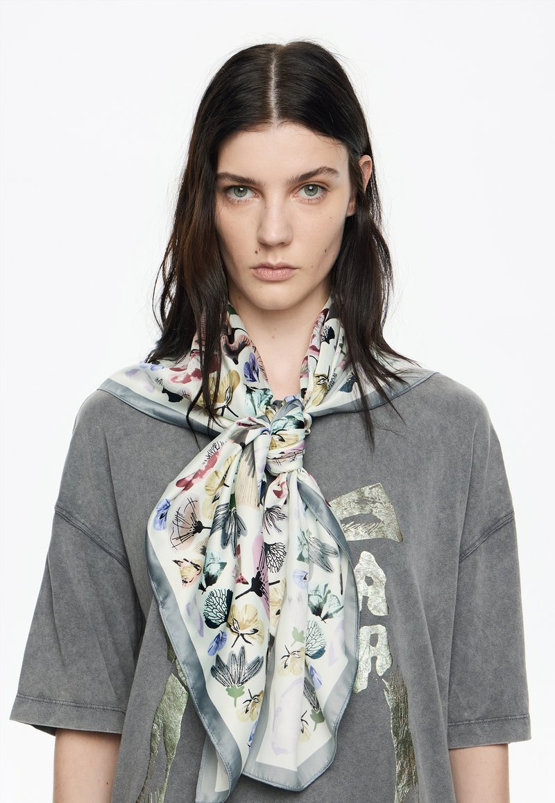 Foulard de seda con un patrón floral en colores pastel, bordeado de gris. Lleva un nudo alrededor del cuello sobre una camiseta gris de gran tamaño.