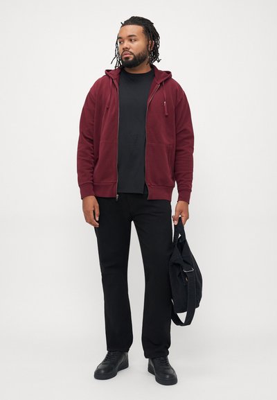 Homem a usar um hoodie burgundy com fecho, camisa preta, calças pretas e sapatilhas pretas, segurando uma bolsa preta, de pé contra um fundo branco.