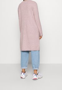 Lång, rosa stickad cardigan med avslappnad passform, matchad med ljusblå jeans med uppvikta manschetter och vita sneakers med färgglada accenter.