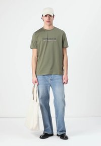 Camiseta verde oliva com o logótipo "Tommy Hilfiger", jeans azul claro, sapatos pretos e uma mala beige; modelo está de pé contra um fundo branco.