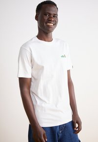 Camiseta de algodón blanca con un pequeño logo verde y rojo en el lado izquierdo del pecho. Cuello redondo clásico y manga corta. Diseño sencillo, ajuste relajado.