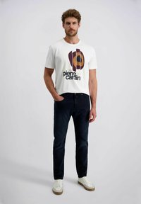 Mann trägt ein weißes T-Shirt mit Grafik von Pierre Cardin, dunkelblaue Jeans und weiße Sneaker und steht vor einem einfarbigen hellen Hintergrund.