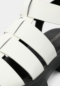 Vita sandaler med breda tvärband av syntetmaterial, med svart sula och en platt, texturerad fotbädd.