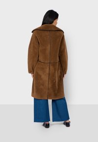 Cappotto in pelliccia artificiale marrone con ampio colletto, dettagli di cuciture verticali e vestibilità relax, abbinato a pantaloni larghi blu e scarpe nere.